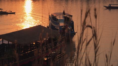 Plano-Exterior-Medio-De-Un-Bote-Que-Viene-Alrededor-De-Otro-Bote-En-Un-Lago-Con-El-Reflejo-Del-Sol-En-La-Hora-Dorada-A-Través-De-Un-Poco-De-Hierba