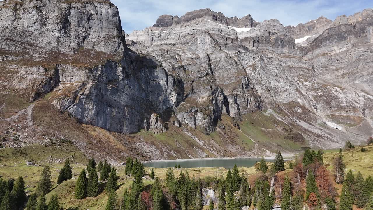 Aerial wide shot of colorful conifer trees and lake with steep mountain range in background. Sunny day in fall season.