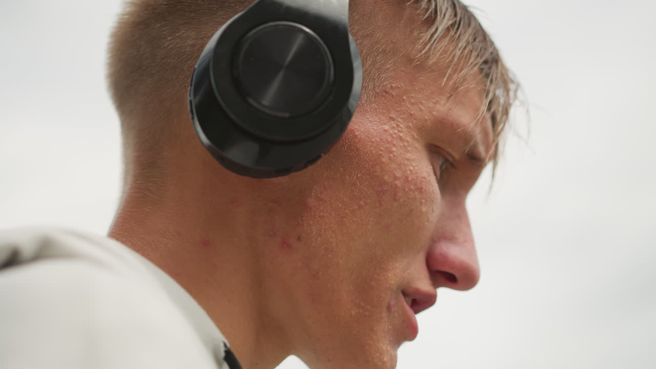 primer plano auriculares gotas caucásico hombre ajustando, almohadilla texturizada, gotas de agua brillantes, horizonte silenciado, expresión sutil, presionar en la oreja con la mano, ambiente de audio portátil, ambiente de llovizna urbana, táctil