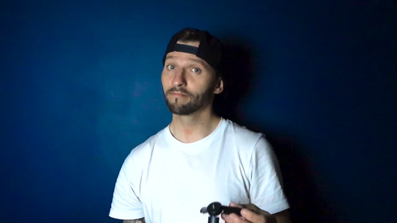 Tattooed man with a white shirt and a black colored reverse cap hat showing and comparing two black tripods that are meant for filming gear. One tripod is small and the other is big. Blue wall at back