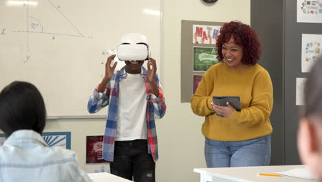 african american boy using vr headset in classroom while female teacher with tablet, at school