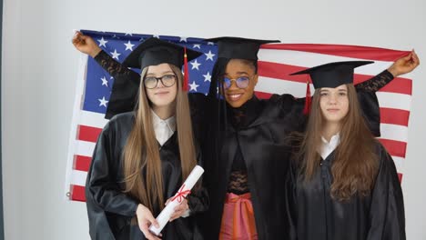 tres jóvenes graduadas felices de diferentes nacionalidades se paran juntas uno al lado del otro y sostienen diplomas y la bandera de los estados unidos detrás de ellas. vista de cerca sobre fondo blanco