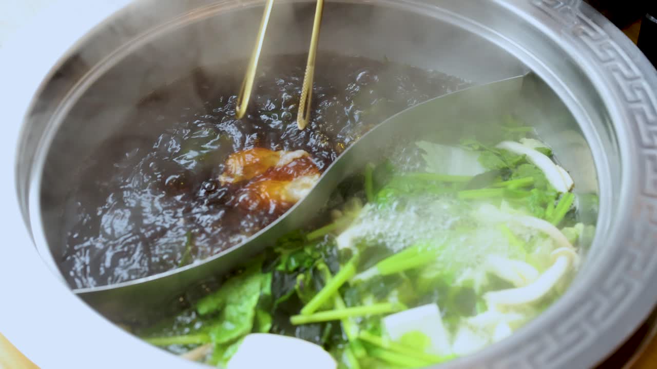 Chopsticks cook sliced meat in steaming dual broth hotpot with fresh vegetables, overhead view
