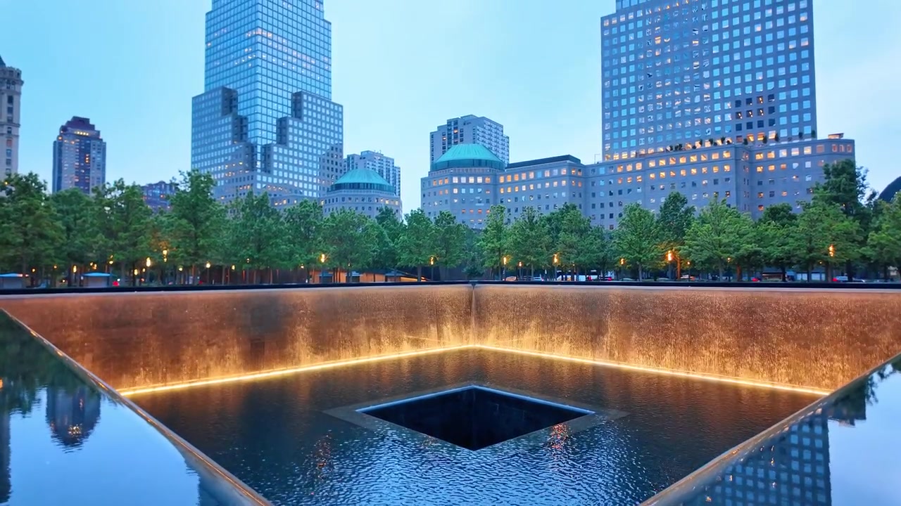 Estanque reflectante y cascada del monumento conmemorativo del 11-S al crepúsculo, Bajo Manhattan, Nueva York. Amplia composición simétrica de muros de cascada iluminados flanqueando el vacío central, torres de la ciudad y árboles al fondo.