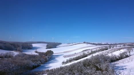 drone flight over snow-covered agricultural landscape on sunny day in winter with wind turbines in distance