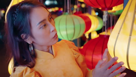 Young-Vietnamese-woman-in-traditional-outfit-looks-and-touches-yellow-lantern-in-Hoi-An,-Vietnam