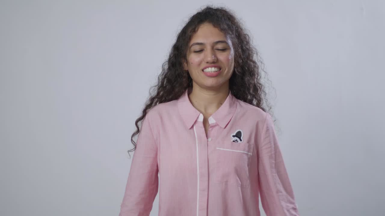 A South Asian woman with curly hair, in a pink shirt, makes a disgruntled or disgusted face and then smiles back again