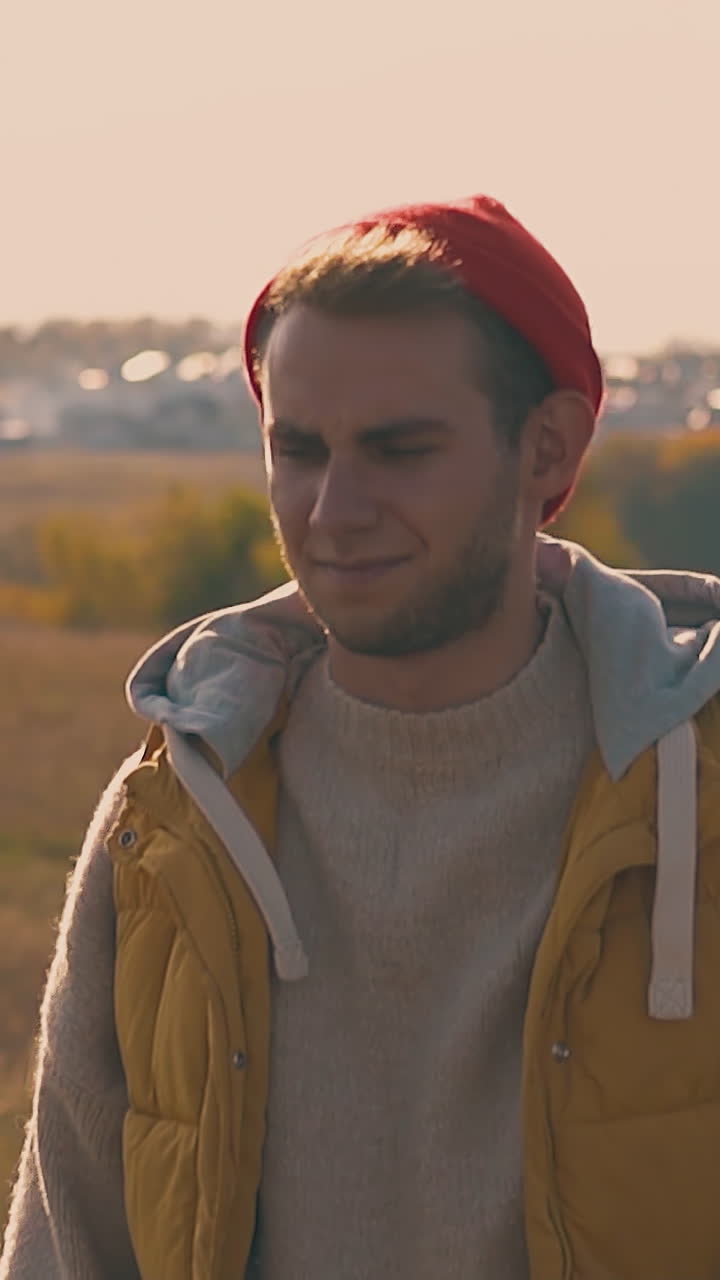 smiling guy fixes orange hat and walks away on yellow meadow against blurry village house silhouettes slow motion