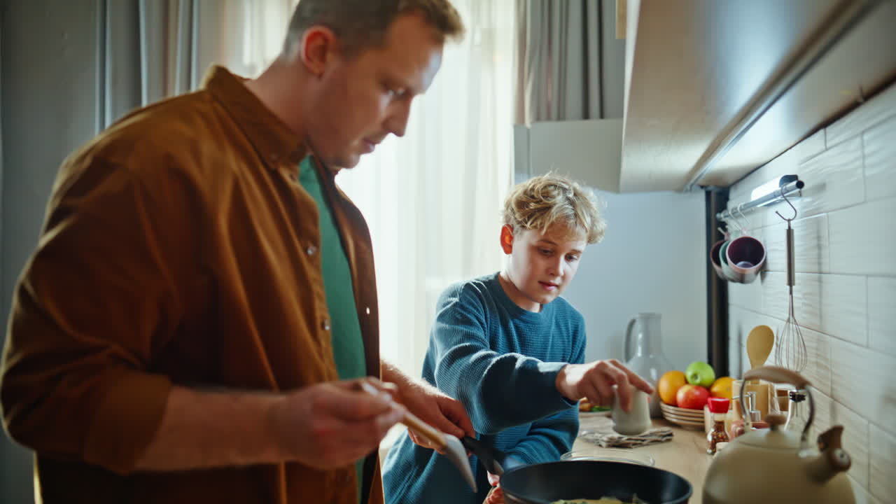 Nahaufnahme: Sohn salzt Bratgericht und hilft jungem Vater beim Zubereiten des Morgenfrühstücks.