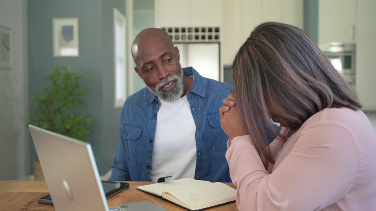 Mature couple sitting at kitchen table reading laptop, pointing notes, smiling and working