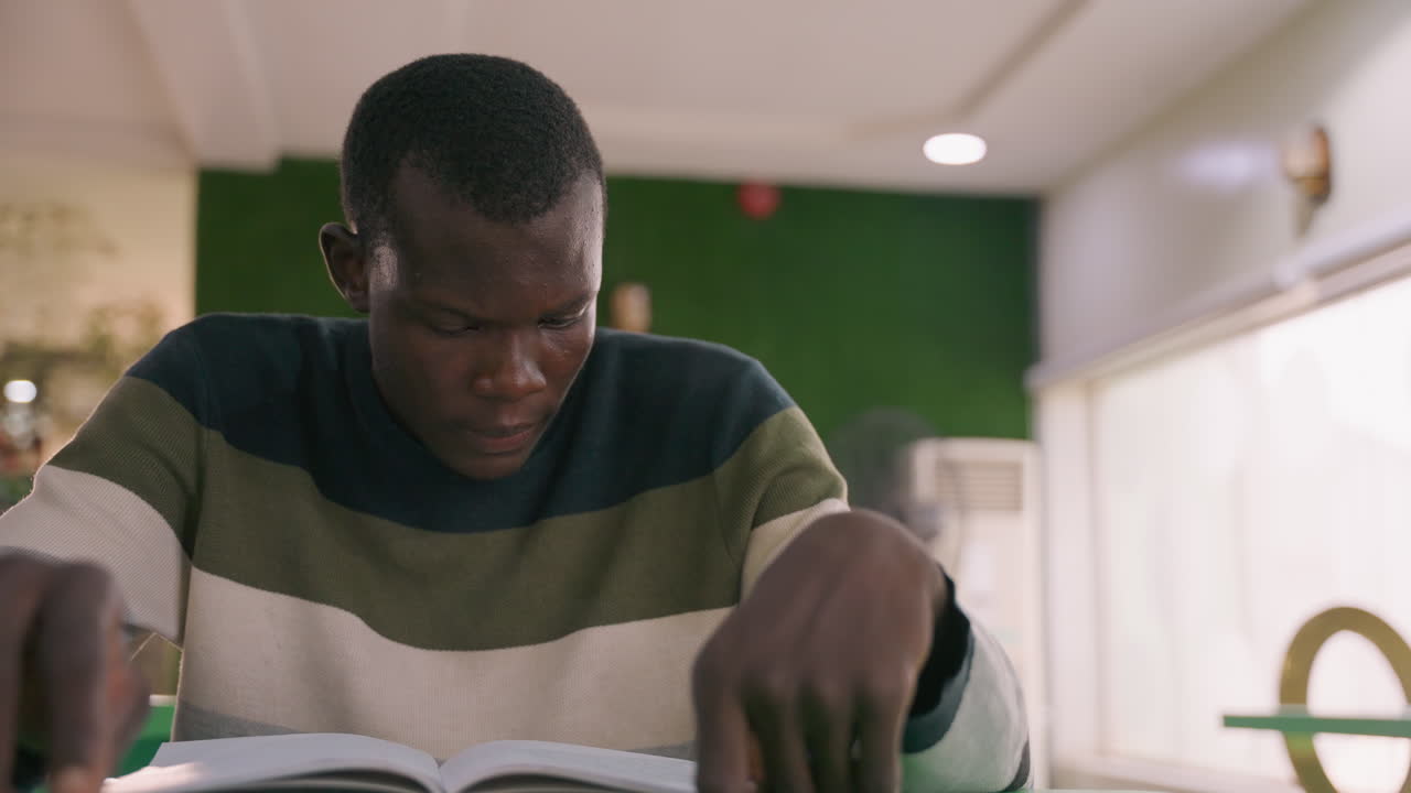 Giovane tranquillamente impegnato in una sessione di lettura al chiuso. Studente si concentra tranquillamente su un libro all'interno di un accogliente caffè. Giovane si rilassa in silenzio mentre è immerso nella lettura in un ambiente caldo da caffè.