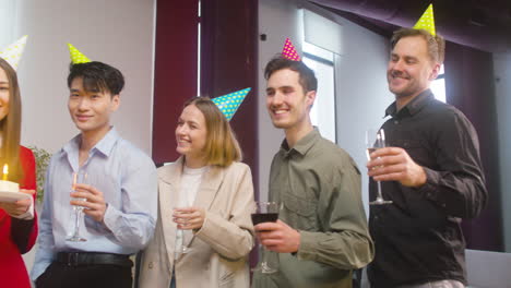 Portrait-Of-A-Woman-Looking-At-The-Camera-And-Holding-A-Birthday-Cake-Surronded-By-Multiethnic-Colleagues-Who-Raising-A-Toast-At-The-Office-Party