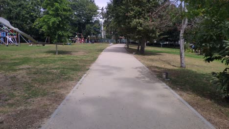 Adultos-Con-Niños-Disfrutando-De-Los-Juegos-Del-Parque-Público-Con-Sus-Jardines-Y-árboles-Una-Soleada-Mañana-De-Verano