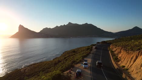 car-driving-along-Hout-Bay-coastline-with-sunset-over-Sentinel-Peak-in-Cape-Town,-aerial