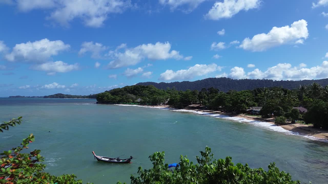 Cinematic aerial drone flight above dense tropical forest in Koh Lanta, Thailand, showing colorful longtail boats anchored in emerald water and revealing paradise beach with palm trees and bungalows