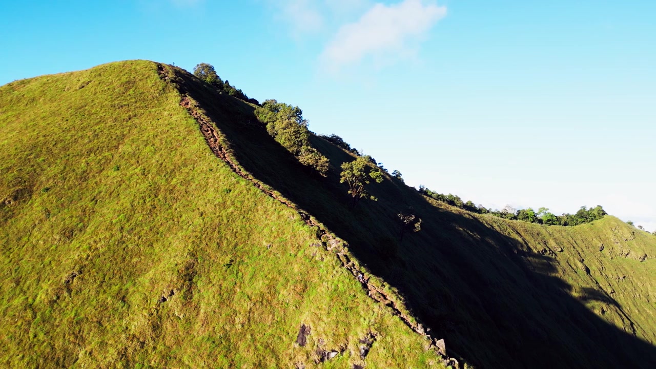 Escursionista di montagna che cammina lungo uno stretto sentiero di cresta circondato da ripidi pendii verdi e un aspro paesaggio montano in Indonesia, riprese aeree con drone.
