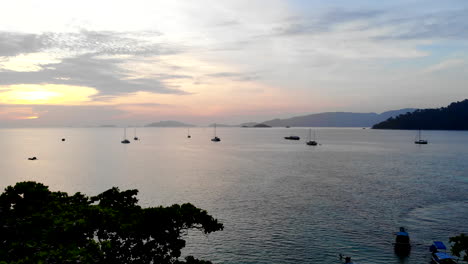 Grupo-De-Personas-En-Una-Playa-Viendo-La-Puesta-De-Sol-Sobre-Barcos-Y-Montañas