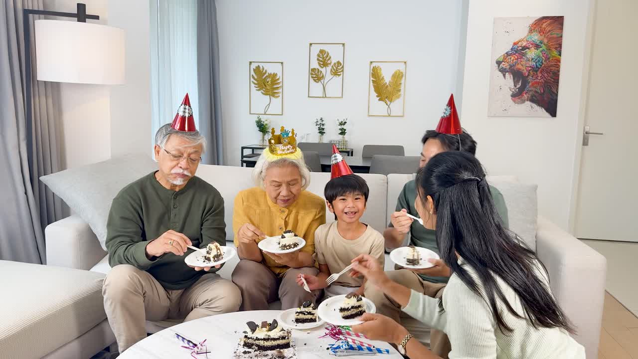 Four generations enjoy birthday cake in a bright living room, sharing smiles and festive hats