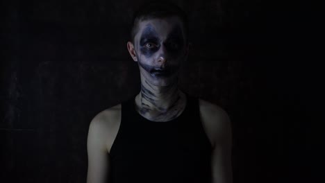 young caucasian man with a scary dark halloween clown makeup costume standing in front of a black background