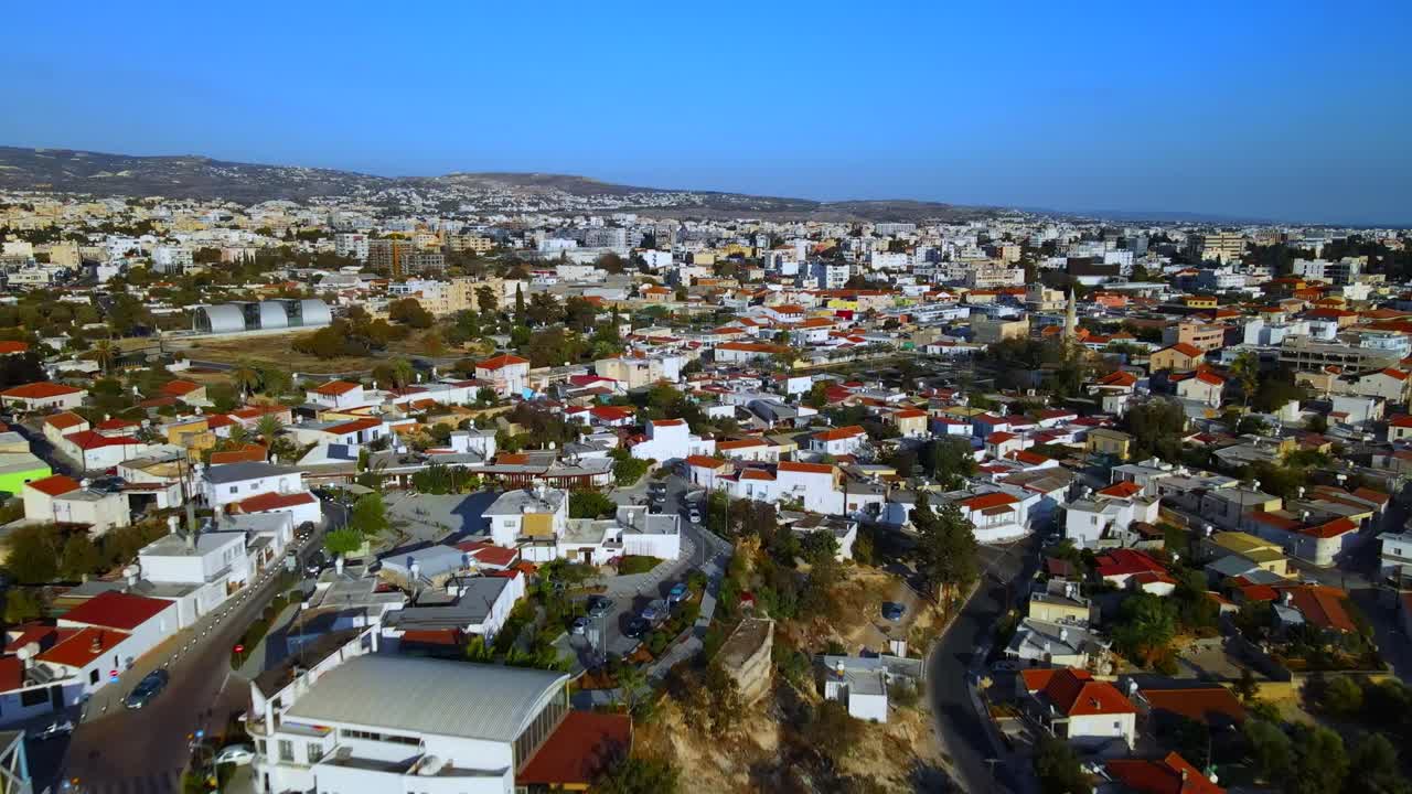 Drone shot of compact Mediterranean neighborhood filled with red-tiled rooftops, winding streets, and white buildings extending across hills in warm daylight
