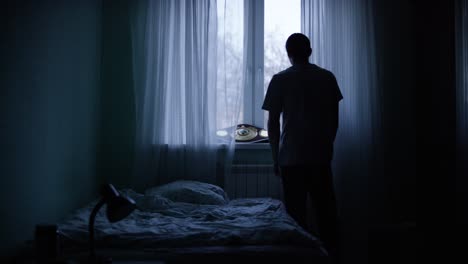 a young man standing by the window in a dark bedroom