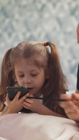 cute little girl with ponytails and happy daddy do homework online with modern smartphone lying on floor in light room at quarantine time