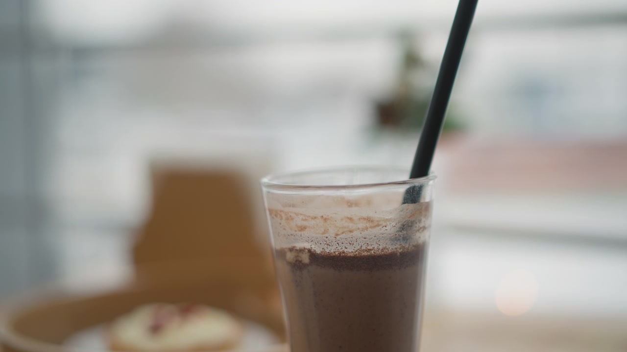 Un alto bicchiere di plastica trasparente riempito con una bevanda fredda color cioccolato, schiuma sottile e piccole bollicine superficiali visibili in cima, una cannuccia nera inclinata all'interno, posizionato su un tavolo di legno con una sedia e una finestra sfocate.