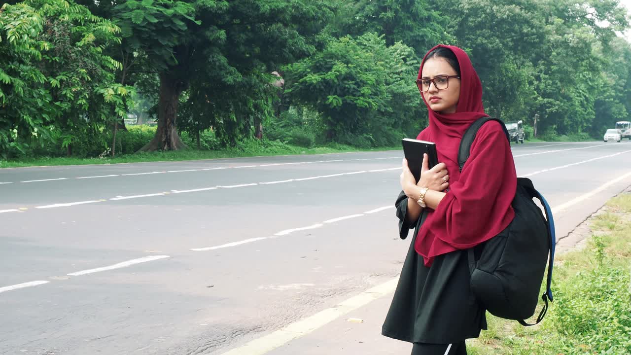 Beautiful serious young Afghan woman in hijab holding file and bag in hand and standing on street near road car and bike passing in the background, charming female in black t shirt