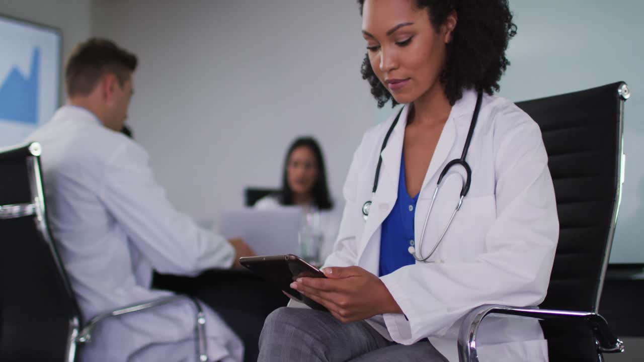 Mixed race female doctor sitting in meeting room using tablet looking to camera smiling