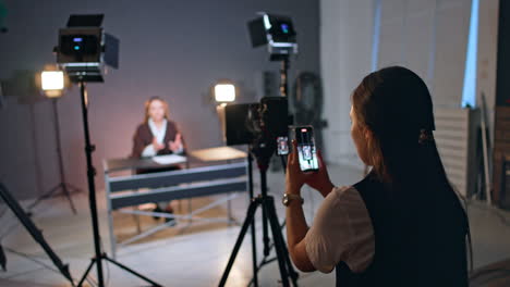 studio footage of a blog content. rear view of a girl filming a female reporter on her phone near the professional camera.