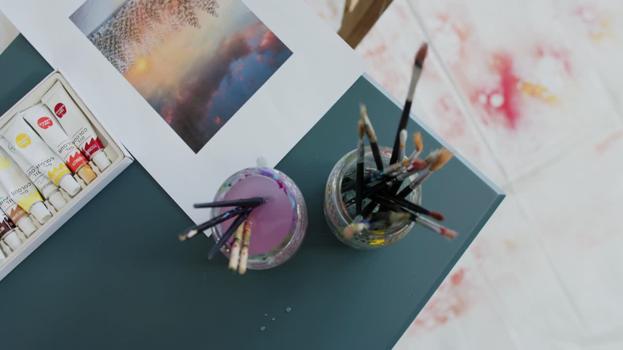 Mão entrando no canto superior esquerdo, pegando pincéis de um pote roxo e enxaguando em uma mesa verde-água para pintar.