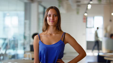 Portrait-Of-Young-Smiling-Businesswoman-Standing-In-Busy-Modern-Open-Plan-Office