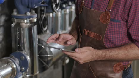 young male brewer wearing a leather apron supervise the process of beer fermentation at a modern brewery factory