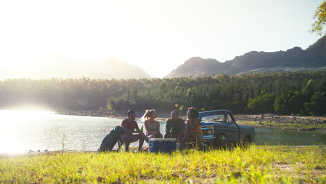 group of friends with backpacks by pick up truck on road trip drinking beer from cooler by lake