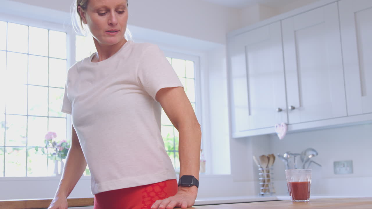 Donna matura in abbigliamento sportivo con una bevanda salutare a casa in cucina, che controlla l'attività sul suo smartwatch