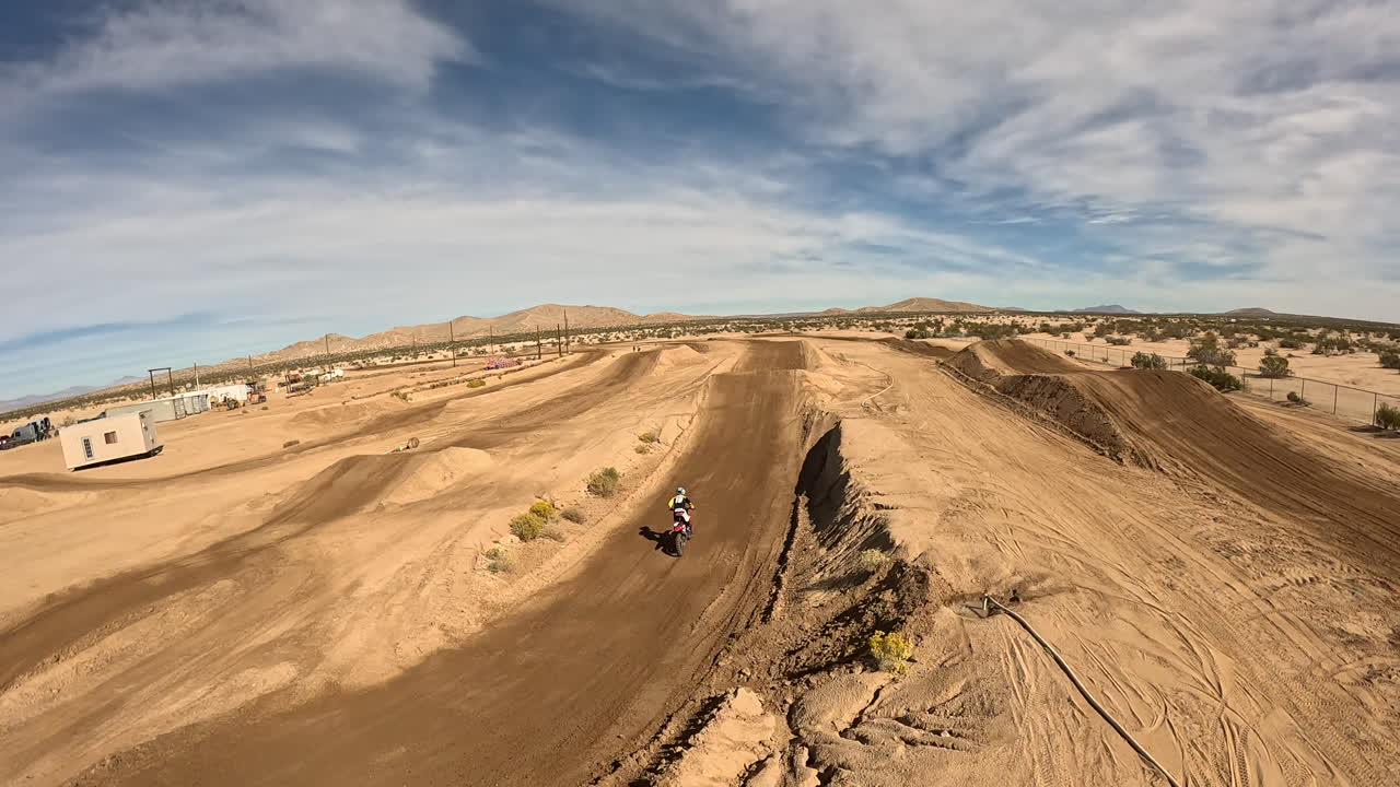 motocicleta dando saltos largos mientras acelera a lo largo de una pista de carreras de tierra seguida por un dron con vista en primera persona
