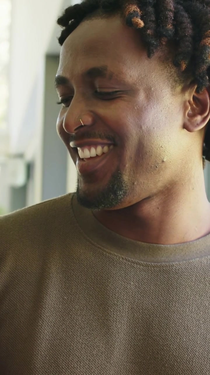 Vertical video: Talking man by window with partner smiling, shifting to laughter in olive sweater