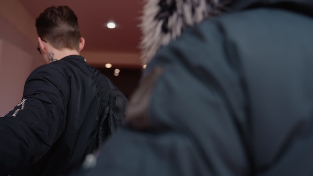 Two guys wearing jackets walk towards glass entrance of wooden building, one carrying black backpack, other with fur hood coat, urban lifestyle moment in daylight as they approach door together