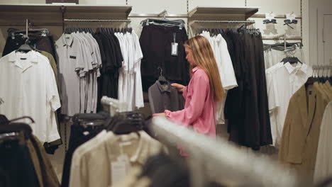 lady dressed in pink outfit and carrying a black bag carefully examines a grey top in a well-arranged clothing store, the store is well-lit with racks of clothes organized in the background