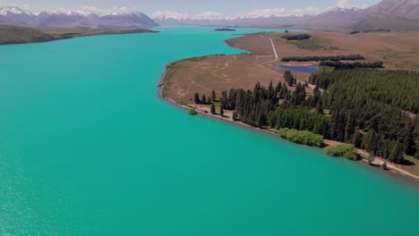 Fliegen-über-Den-Tekapo-See-Mit-Schneebedeckten-Bergen-Und-Wald-Im-Hintergrund