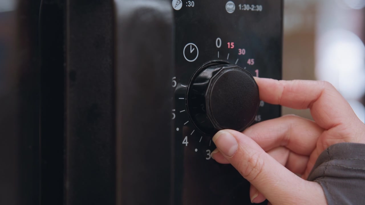 Quadrante del timer del microonde con numeri rossi luminosi, mentre una mano imposta il conto alla rovescia ruotando una manopola testurizzata verso i quindici minuti. Pannello di controllo nero lucido. Atmosfera da cucina commerciale con preparazione concentrata e precisa.