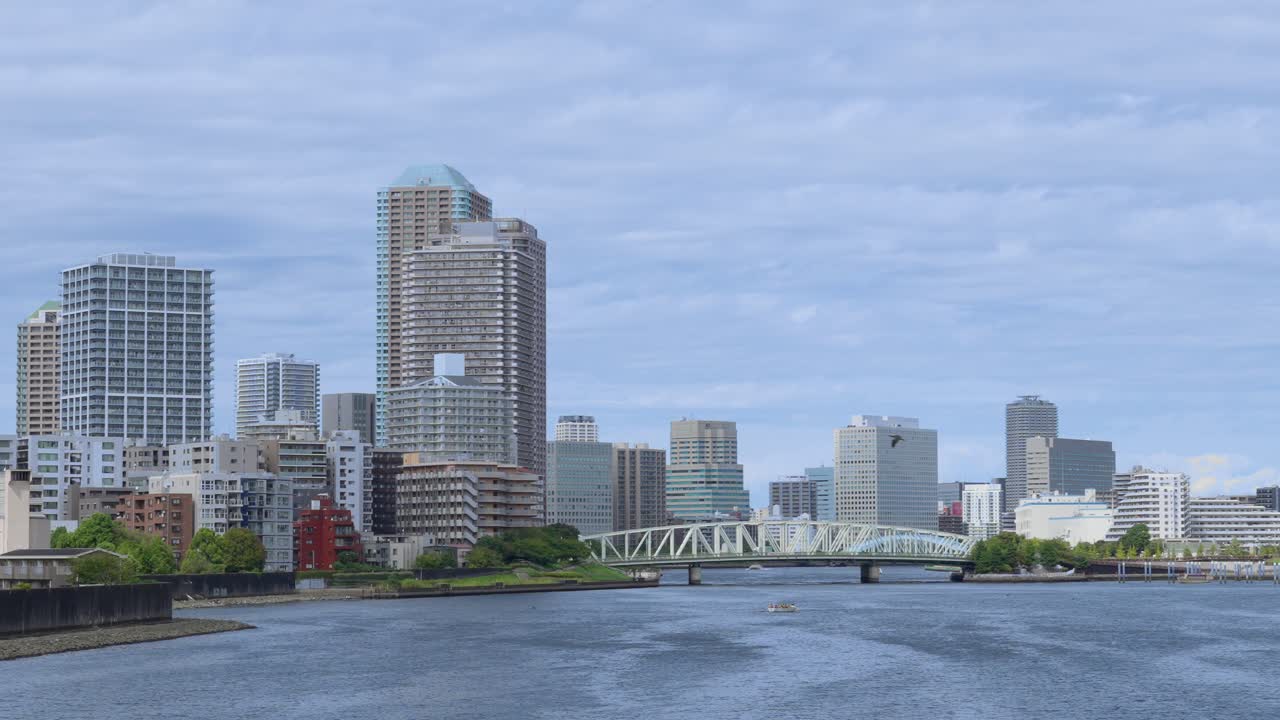 A wide shot of the Tokyo cityscape from a river, showing a mix of modern skyscrapers and a bridge