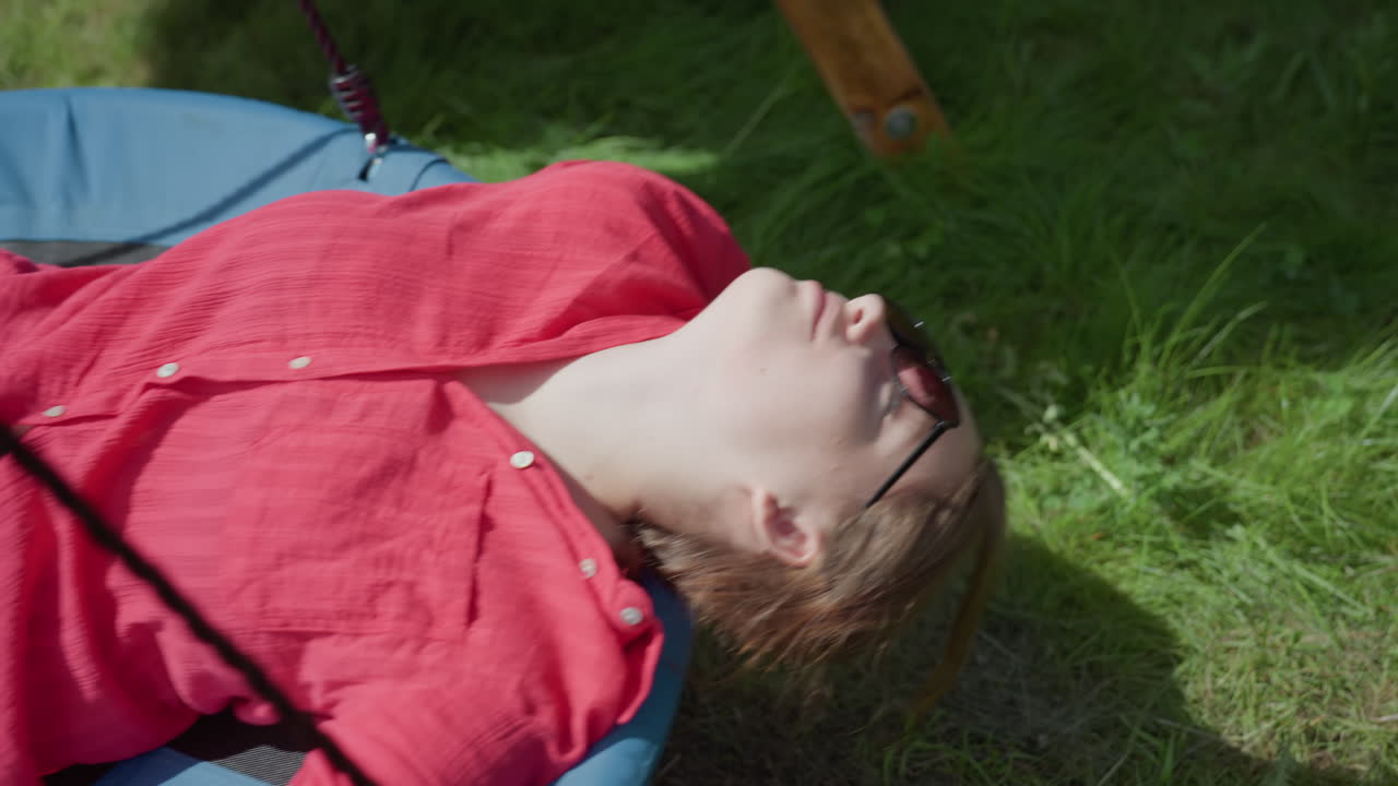 Woman in red shirt and sunglasses lays back on blue swing, soaking up sun in quiet grassy backyard, eyes closed, arms spread, fully relaxed in peaceful outdoor environment with warm natural light