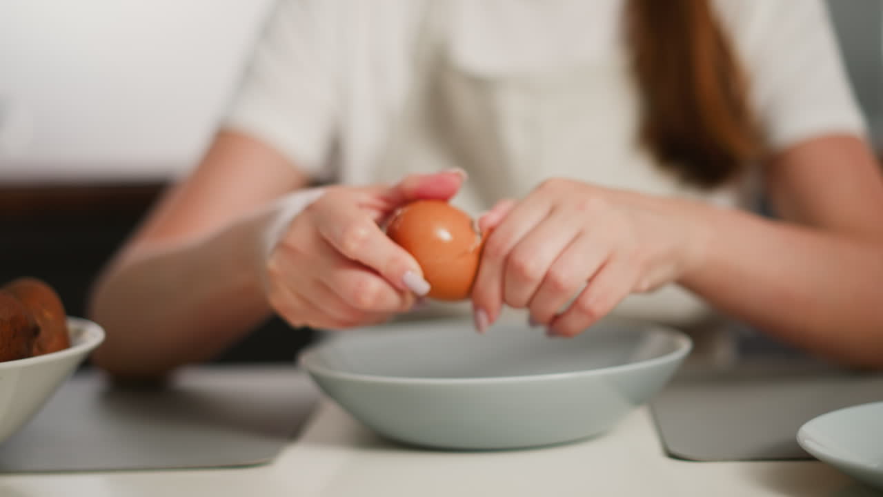 Close up of woman cracking egg shell on glass plate then peeling shell from boiled egg over bowl at kitchen table, wearing apron with potatoes and dishes in background, showing cooking prep