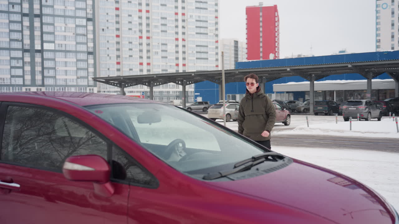 Young man in winter jacket walking toward parked car ready to enter with snow covering ground, residential high-rise buildings and multiple parked vehicles in background during cold winter day
