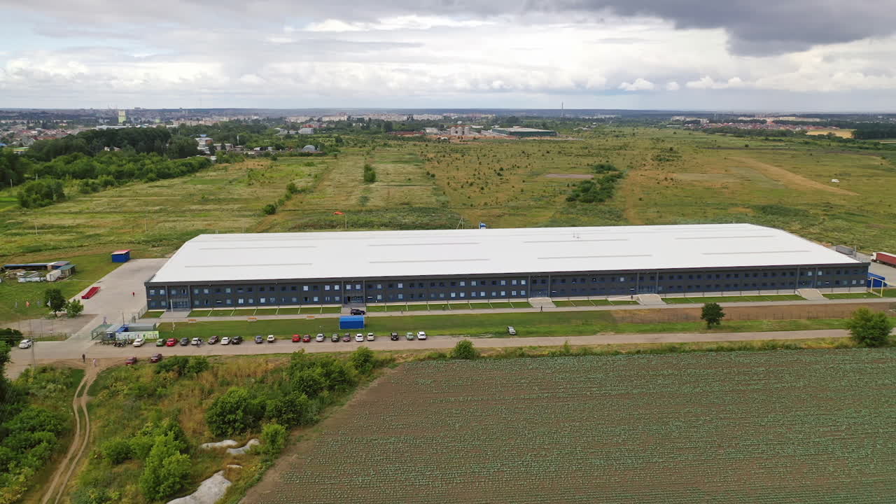 Aerial panorama of a modern plant. Exterior of a large commercial warehouse situated among green fields. Camera moves forward.