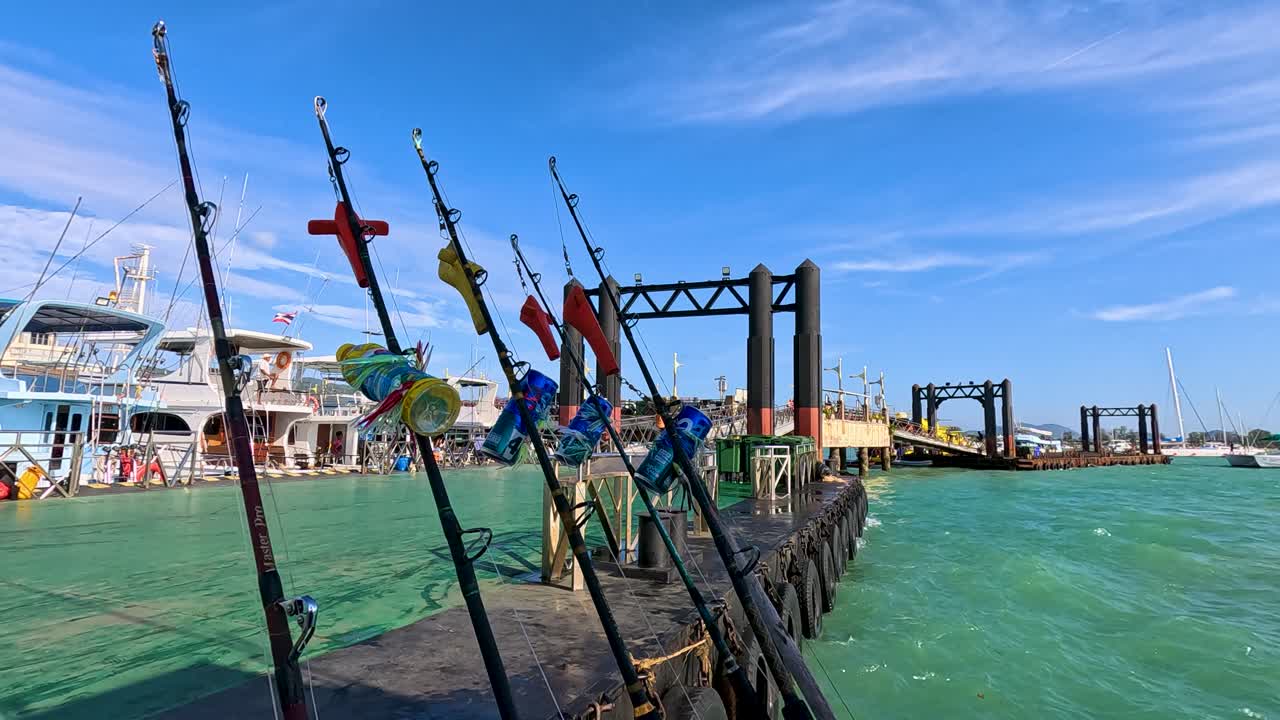 Fishing rods line a jetty under clear skies at Rawai Beach, Phuket. Vibrant colors and calm waters create a serene atmosphere