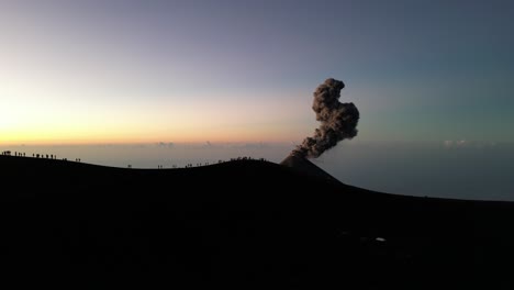 Drone-view-in-Guatemala-flying-over-a-volcano-crater-with-an-erupting-volcano-at-sunrise-and-shadow-of-people-on-the-rim