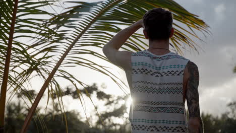 View-Behind-Male-Wearing-Tank-Top-Jumping-Up-Beside-Palm-Tree-To-Seek-Out-Sunset-In-Punta-Cana