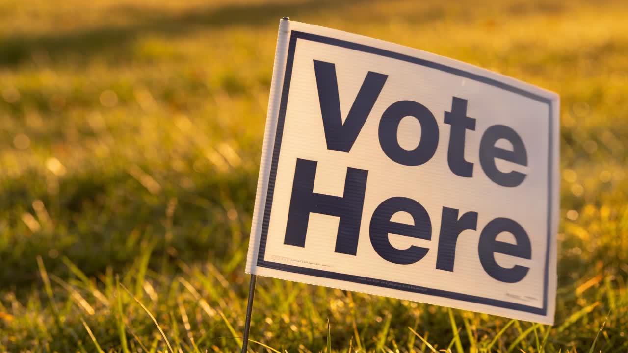 Pushing camera in while yard sign reading Vote Here swaying on wire stake in grassy lawn